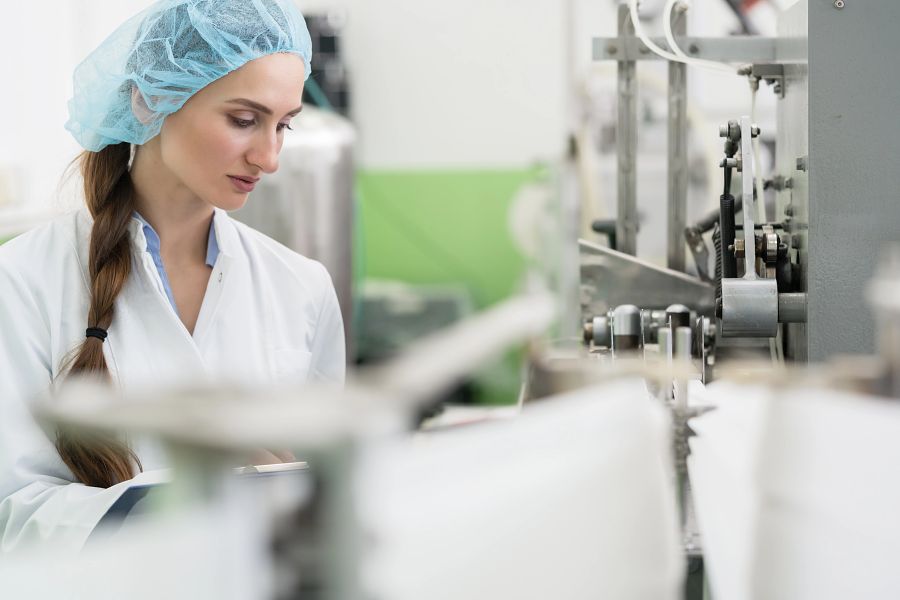 Happy female employee wearing protective headwear and white lab coat while working as a manufacturing engineer in a contemporary cosmetics factory