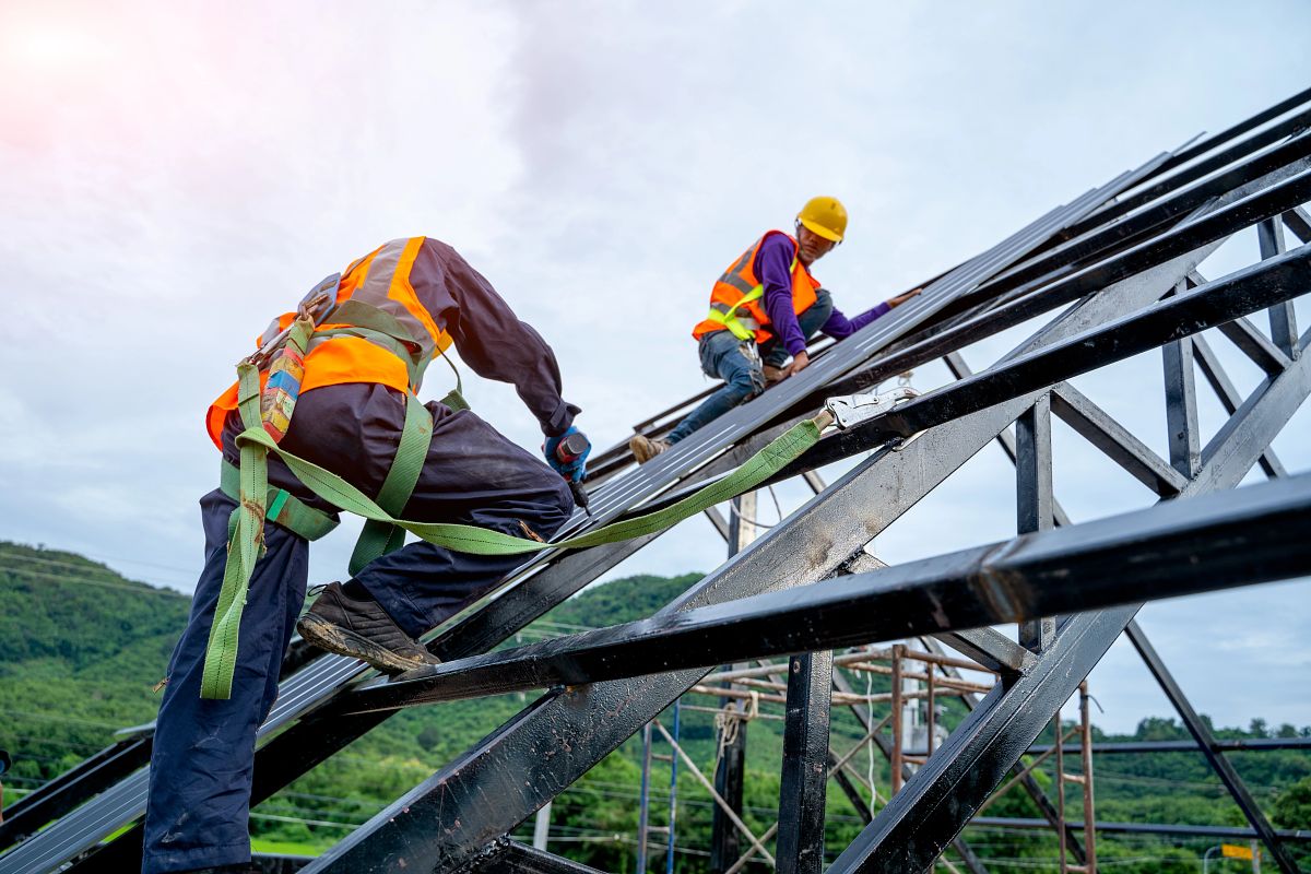 Roofer worker in protective uniform wear and gloves, using air or pneumatic nail gun and installing asphalt shingle on top of the new roof