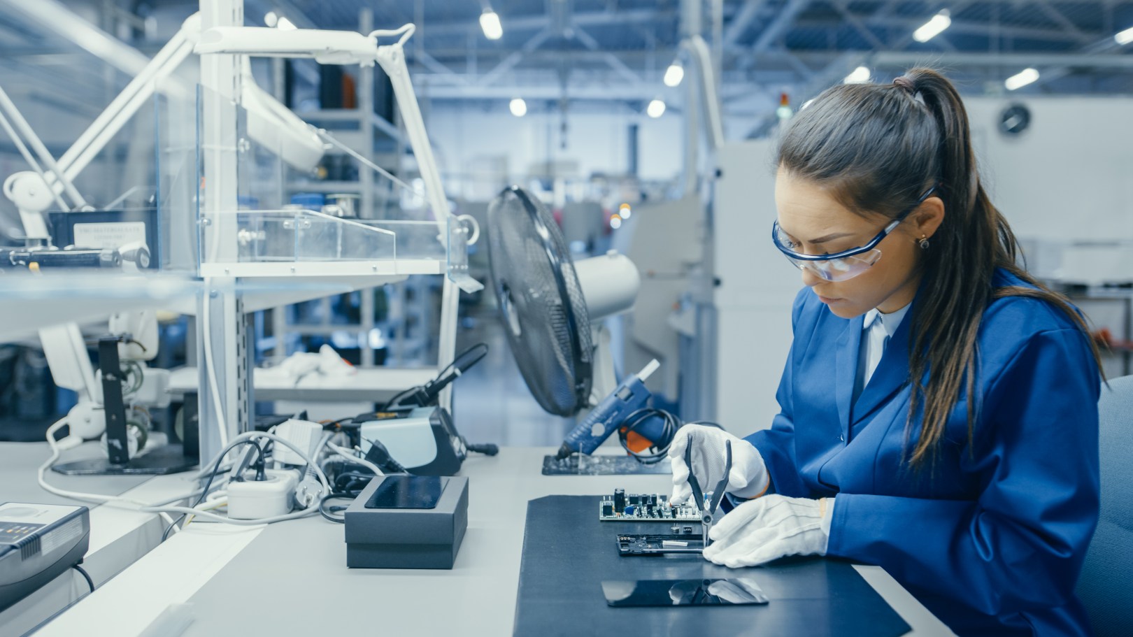A person in safety glasses and a lab coat working on a mobile phone with small pliers.
